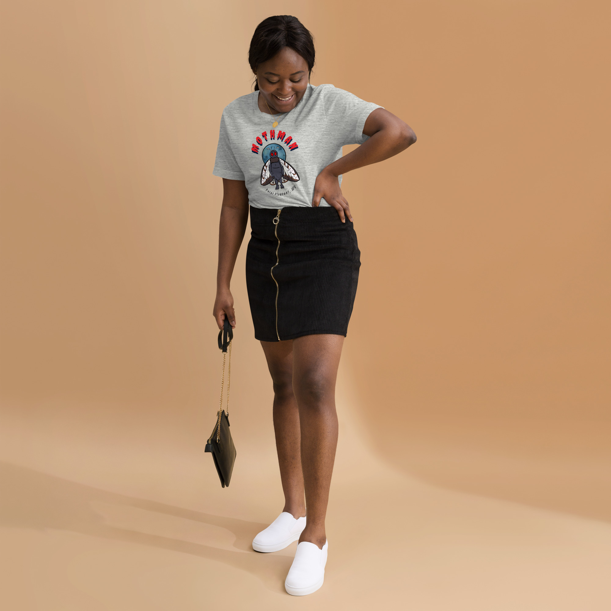 Woman wearing grey Mothman graphic tee, black zip-front corduroy mini skirt, and white slip-on shoes