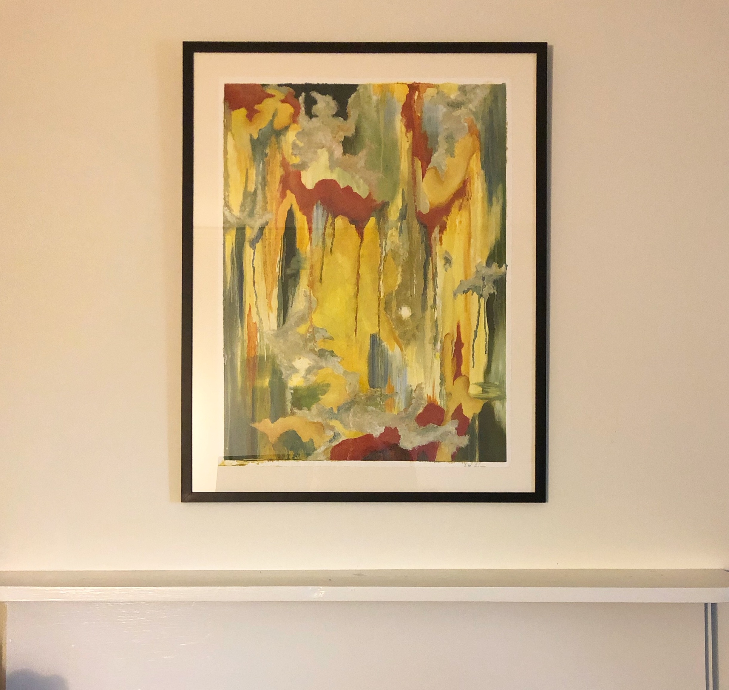 Abstract painting with vertical drips of red and yellow in a black frame hanging on a wall above a shelf.